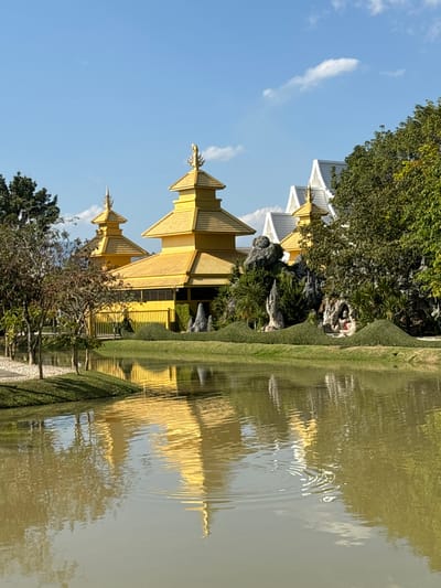 A golden, multi-tiered structure with ornate roof peaks stands alongside a body of water, possibly a pond or lake. Its reflection shimmers on the water's surface, creating a mirror image. Lush green landscaping flanks the structure and water, including manicured hedges and trees. A rocky outcrop with foliage sits near the water's edge. The sky is blue with scattered clouds, enhancing the brightness of the gold.