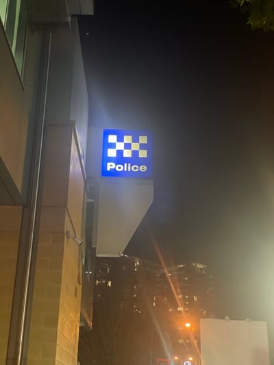 An illuminated "Police" sign is mounted on the exterior of a building at night in Fortitude Valley, Australia. The sign features a blue background with a yellow and blue checkered emblem above the word "Police" in white lettering. The building appears to be made of light-colored brick. A security camera is visible mounted on the brick wall below the sign. In the background, trees and illuminated apartment buildings are visible, indicating an urban environment. The scene is lit by artificial lights, including the police sign and streetlights, creating a somewhat hazy atmosphere due to the night and potential fog or dust.