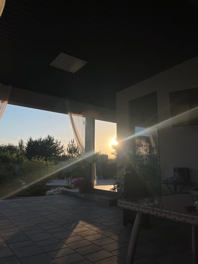 This media file captures a view of an outdoor patio area during sunset, with the sun's rays creating lens flare across the image. The scene is oriented vertically. On the left side, a dark, covered porch or overhang occupies a significant portion, with a rectangular light fixture visible on its ceiling. Beyond the overhang, a bright opening reveals the sky and surrounding environment.
The setting appears to be a residential patio with paving stones. To the right of the opening, a white table with a woven surface and chairs are partially visible, suggesting an outdoor seating area. Further into the background, lush green trees and bushes are seen, with some flowering plants in pots adding color. The sky is clear and light, indicative of a warm, late afternoon or early evening. The sun, low on the horizon, is the primary light source, casting long shadows and illuminating the scene with a golden hue.
No people are visible in the image. The overall mood is peaceful and serene, enhanced by the warm lighting of the sunset. The location context provided is Novooleksandrivka, Ukraine, which aligns with the residential patio setting. There is no visible text in the image.