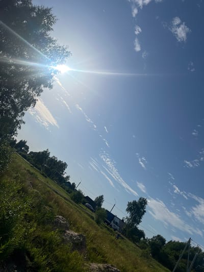 This is a tilted, eye-level, outdoor shot of a rural landscape on a sunny day. The sun is bright and positioned in the upper left quadrant, casting lens flare and creating a strong light source. The sky is a clear blue with scattered white clouds, some of which are thin and wispy, while others are more cumulus-like.
The foreground is dominated by a grassy slope covered in various types of vegetation, including tall grasses and bushes, with some rocky or dirt formations visible. The terrain appears uneven, with a noticeable incline.
In the midground and background, trees are present, including what appear to be birch trees. The presence of these trees, along with the style of the buildings visible, suggests a rural or semi-rural setting, consistent with the location of Ilyanskiy selskiy Sovet, Belarus. A road or path winds through the landscape, and several vehicles, including a white truck and a dark-colored car, are parked or driving on it. A few structures, possibly houses or outbuildings, are visible among the trees. Power lines and poles are also present, indicating the presence of infrastructure. One individual can be seen walking on the grassy slope in the lower midground. The overall impression is one of a warm, sunny day in a countryside environment.