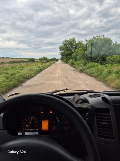This image depicts a view from inside a vehicle moving along a rural road. The perspective is from the driver's seat, where a steering wheel and dashboard with illuminated instruments (including a speedometer, warning lights, and digital odometer) are visible. Outside, the road is a narrow, seemingly older concrete or asphalt surface, flanked by greenery. The sky is overcast with cloudy conditions. The location is unknown.