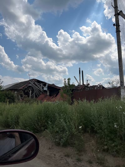 The image presents a landscape in Staryi Saltiv, Ukraine, characterized by significant structural damage and overgrown vegetation, viewed from the perspective of a vehicle. The foreground features a dirt path and dense, green, waist-high undergrowth. In the lower left, a maroon vehicle's side mirror is visible, reflecting a portion of a person wearing a white shirt.
Centrally, two heavily damaged structures are prominent. The structure on the left, likely a house, has a collapsed roof with exposed and broken wooden rafters and beams, indicating severe destruction. A small, disused antenna is still attached to the remnants of its dark roof. To its right, behind a reddish-brown corrugated metal fence or wall, a pile of rubble includes several dark, vertical pipe-like objects, consistent with shattered chimneys or ventilation shafts.
On the far right, a concrete utility pole stands upright, supporting multiple wires. A white triangular sign with black numbers "92" is affixed to the pole. The sky above is blue with large, scattered white cumulus clouds, and sunlight is visible, casting faint rays. No other individuals or active human interactions are present in the primary scene.