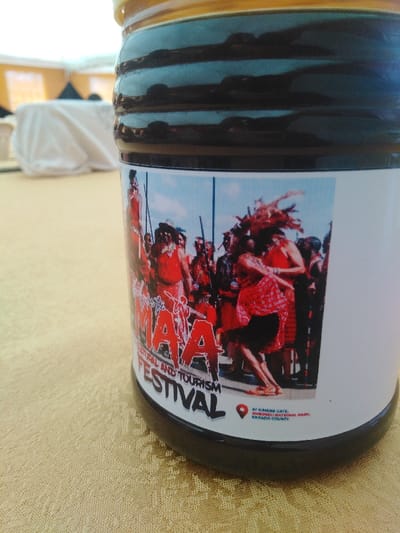 The image features a dark, cylindrical jar with a golden-yellow screw-top lid, positioned on a light brown, subtly patterned tablecloth. The jar contains a dark, viscous substance. A white label is adhered to the jar's lower section, displaying a colorful photograph and text.
The photograph on the label depicts an outdoor cultural event under a blue sky, featuring a group of approximately ten individuals. Most figures are dressed in red traditional garments, some with elaborate headwear and carrying staffs or spears. One central figure on the right is captured in mid-dance, with hair or a headdress in dynamic motion.
Visible text on the label reads: "MAA CULTURAL AND TOURISM FESTIVAL," with a red map pin icon followed by the location: "AT KIMANANA GATE, AMBOSELI NATIONAL PARK, KAJIADO COUNTY."
The background behind the jar is blurred, showing white-draped tables within an indoor setting. The image itself provides no visual information confirming a location in Sigona ward, Kenya.