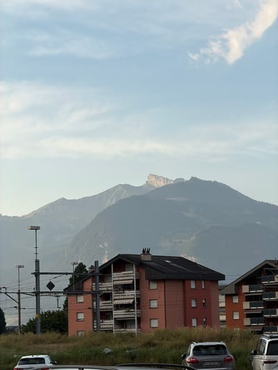 This media file depicts a mountainous landscape with a residential area and a train track in the foreground. The scene is captured at an angle, likely from a vehicle in motion, with the sky on the left and the ground on the right. 

The subjects include several multi-story buildings with a reddish-brown facade and balconies, some of which appear to be apartments or hotels. A prominent train track with electrical overhead lines runs parallel to the buildings. In the far distance, a large, hazy mountain range dominates the background, with its peaks fading into the atmospheric perspective. The sky is a pale blue with wispy clouds.

The activity taking place is not explicit, but the presence of the train track and the slight blur suggest movement, possibly a train passing or the camera being on a moving vehicle. There are no people visible.

Notable details include the architectural style of the buildings, which appear to be of European design. The time of day seems to be either late afternoon or early morning, given the soft light and the hazy mountains. The weather appears to be clear but with some atmospheric haze. The license plate on the grey car parked near the buildings reads "VS 603969". The location context provided is Monthey, Switzerland, which aligns with the mountainous scenery and architectural cues. There are no strong emotions conveyed, but the overall impression is one of a serene, possibly rural or suburban, mountainous setting.