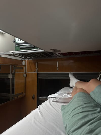The image shows a person's legs and feet resting on a bed within a train compartment. The compartment features wooden paneling, metal shelving, and what appears to be luggage stowed above. The overall impression is that of a confined sleeping space. There is no indication of the location being in Bijelo Polje, Montenegro.
