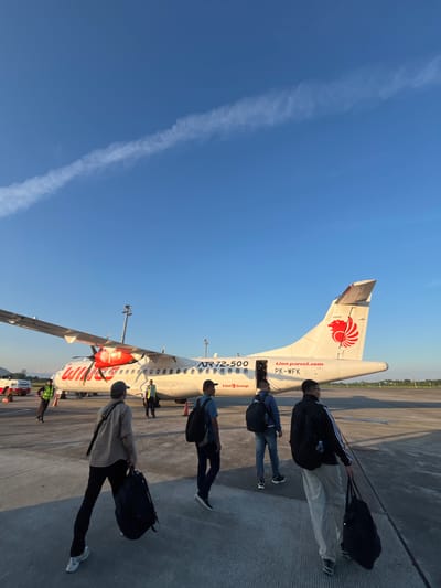 The image shows passengers boarding a Lion Air ATR 72-500 aircraft, registered as PK-WFK, on an airport apron. The blue sky is visible, with a single contrail running vertically. A line of passengers is walking towards the aircraft entrance, each carrying luggage. Ground crew members are present near the aircraft. The location appears to be Baji Mangngai, Indonesia, based on the airline and aircraft registration. The scene conveys a typical moment of air travel during boarding.