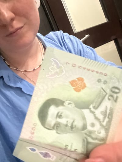 A person is holding a 20 Thai Baht note. The note features a portrait of King Rama X. The scene appears to be indoors, with a blue shirt and a pearl necklace also visible. The location is not discernible from the image.