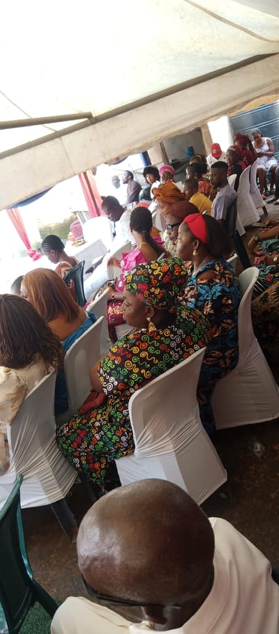 The image depicts a crowded gathering under a white canopy in Udi, Nigeria. The perspective is from behind a bald man's head in the extreme foreground, looking towards rows of seated individuals. Attendees, predominantly adults, occupy white plastic chairs covered with white fabric, arranged in an event setup.
In the mid-ground, a woman wearing a black dress with vibrant multi-colored swirl patterns and a matching headwrap sits on the right, adorned with large, golden, triangular earrings. Next to her, a woman in a blue patterned top and a woman with brown hair are visible. Further back, a woman with braided hair wears a pink top. Various other individuals, both men and women, are seated in the background, dressed in a mix of traditional and casual attire, including patterned fabrics, shirts, and headwraps. One woman in the far background on the right is seen looking at a mobile phone.
The environment suggests an outdoor or semi-outdoor event, with a white tent structure supported by metal beams providing overhead cover. Red and blue drapery is visible in the background, hinting at decorative elements. The ground beneath the chairs appears to be a dark, earthy surface. A partial view of a building structure is discernable beyond the tent in the upper right background.