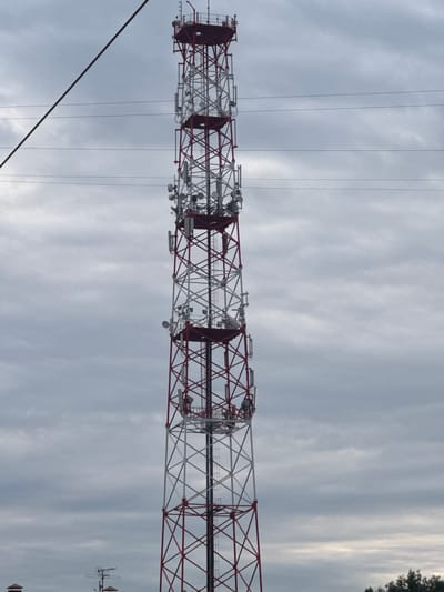 The image shows a tall, red and white lattice telecommunications tower against a cloudy sky. The tower is equipped with numerous antennas and satellite dishes at various levels. Several horizontal power lines are visible crisscrossing in front of the tower and the sky. In the lower left portion of the image, a small portion of another structure with a peaked roof and some antennas is visible, along with the tops of some trees on the right. The scene appears to be an outdoor, industrial or infrastructure area.