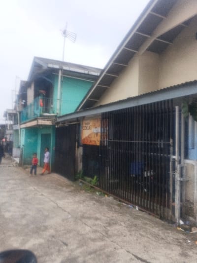 The scene depicts an outdoor urban environment in the city of Morrumbala, Mozambique, under an overcast sky. A paved path or road occupies the foreground and recedes into the background.

On the left, a two-story building painted turquoise features a second-floor balcony with a railing. A red object hangs from this railing. An outdoor television antenna is affixed to the roof of this building. At ground level, a doorway is visible. Two individuals are present on the paved surface in front of this building: a young child in a red long-sleeved shirt and dark pants stands barefoot, partially facing away from the camera. Beside the doorway, an individual in a white top and orange pants also stands barefoot, facing partially towards the camera. Further down the path on the left, additional blurry figures and structures are discernible.

On the right, a building with a light-colored (possibly beige) upper facade and a prominent sloped roof is largely obscured at ground level by a tall, dark metal fence composed of vertical bars. An orange and brown sign, displaying the text "COKLAT PASTI N", is affixed to the fence. Behind the fence, dark, indistinct shapes suggest parked objects, possibly a motorcycle. Litter and debris are scattered on the ground adjacent to the fence line.

The ground surface is concrete or asphalt, showing cracks and general wear. A dark, out-of-focus object, possibly part of a vehicle, is visible in the bottom left corner of the frame.