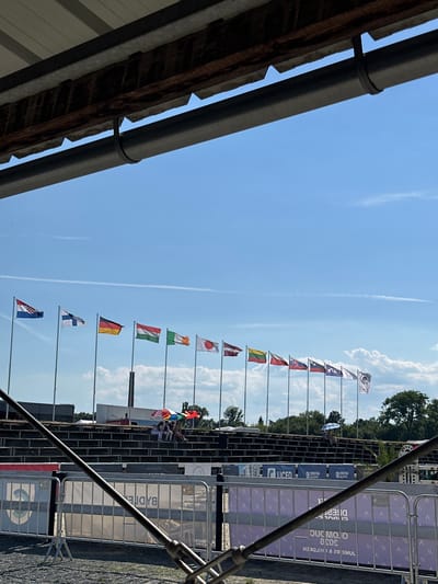 The image captures an outdoor scene with a clear blue sky dotted with wispy clouds. In the foreground, a dark metal pipe runs diagonally across the frame, partially obscuring the view. Behind it, a row of metal fences with a white mesh panel and a blue and white circular logo can be seen. Beyond the fences are tiered stadium seating made of dark wood or metal, with several people seated on them, some holding colorful umbrellas. A line of national flags on poles stretches across the horizon against the sky. These flags represent various countries including Croatia, Finland, Germany, Hungary, Ireland, Japan, Latvia, Lithuania, Poland, Slovakia, and possibly others further to the right. The flags are attached to tall poles and are gently waving in the breeze. In the background, trees and some buildings are visible. The overall impression is of an outdoor event or competition, possibly a sporting event given the stadium seating and flags. The location is not explicitly identifiable as Olomouc, Czechia, based solely on the visual information in the image.
