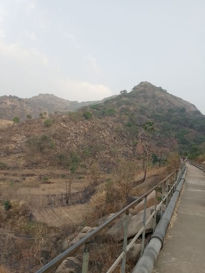 Hills dominate the background, sparsely vegetated with trees and rocks. A concrete pathway stretches into the distance, lined with a metal railing and a large pipe running along its side. Barren, terraced fields and scrubby vegetation fill the area between the path and the hills. A hazy sky creates a muted, overcast light.