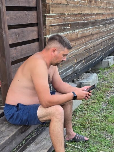 A shirtless man in blue shorts and a black sandal sits on a wooden bench, looking at his phone. He has short, dark hair and a light complexion. Behind him is a rustic wooden structure with horizontal planks and a rough, textured finish. The ground is covered in dry grass and small stones. The scene appears to be outdoors, possibly in a rural or semi-rural setting.