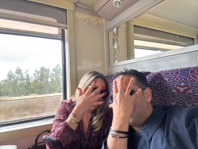 The image depicts two individuals inside a train carriage, reportedly in the city of Sidi Yahya El Gharb, Morocco. A female with light-colored hair and a dark red patterned garment is on the left, her right hand with red nail polish, gold bracelets, and a gold ring raised to obscure her face. She wears dark sunglasses. A black smartphone is visible near her left. A male with dark hair and a dark blue collared shirt is on the right, his left hand with black bracelets and a silver anchor charm raised to obscure his face. He wears dark sunglasses. Both individuals are looking towards the camera, intentionally covering their faces.
The train interior features upholstered seats with a purple, blue, and white geometric pattern. A partial seat number "46" is visible on a headrest. Light-colored wall panels and a ceiling light fixture are visible. Above the male, a decorative silver-toned panel with a patterned cut-out design is mounted. A large rectangular window on the left side displays an exterior scene composed of numerous green trees under an overcast sky, with a blurry light brown area below the tree line.