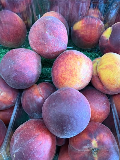 A close-up, overhead shot displays a collection of ripe peaches arranged in clear plastic containers on a green, artificial turf surface. The peaches vary in color, exhibiting shades of red, pink, orange, and yellow. Some peaches show a dusty white bloom on their skin. The scene appears to be set in a market or grocery store in Kadima-Zoran, Israel, given the context provided. There are no people visible in the image.
