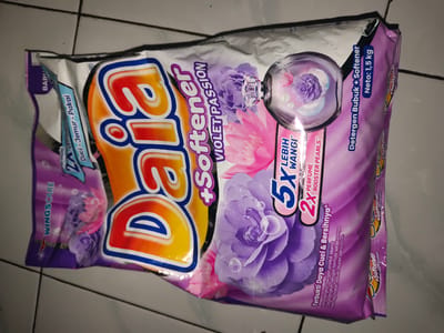 This media file shows a close-up of a package of "Daia" brand laundry detergent with added softener. The packaging is predominantly purple and white, featuring large, colorful graphics of flowers, specifically roses and what appear to be peonies, with water droplets. A prominent circular graphic depicts a glass sphere containing flowers and bubbles, suggesting a refreshing scent or effect. The brand name "Daia" is displayed in large, stylized orange and yellow letters with a blue outline.

Key text visible on the packaging includes "Daia +Softener VIOLET PASSION," "Detergen Bubuk + Softener," and "Neto: 1,5 kg," indicating the product is a powdered detergent combined with softener, featuring a "Violet Passion" scent, and has a net weight of 1.5 kilograms. Other phrases like "5X LEBIH WANGI" (5X More Fragrant) and "2X PERFUME BOOSTER PEARLS" highlight its scent-related benefits. The phrase "Terbukti Daya Cuci & Bersihnya" (Proven Washing Power & Cleanliness) emphasizes its cleaning efficacy.

The package is lying flat on a tiled surface, likely a floor, as indicated by the grid pattern of light-colored tiles. The lighting appears to be artificial, casting some shadows and giving the image a slightly dim, indoor feel. The overall impression is a product shot focused on the packaging's design and information, typical of e-commerce or advertising materials. No people are visible in the image. The context suggests the product is intended for household use in Indonesia, given the brand name and language on the packaging.