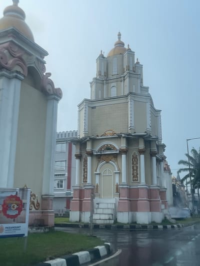 The image shows a building with a grand, ornate architectural style, featuring a prominent golden dome and intricate decorative elements. The building's facade is painted in light colors, with accents of pink and gold. The architecture suggests a cultural or entertainment venue, possibly a theater or a themed attraction. In the foreground, a wet asphalt road with black and white striped curbs indicates recent rainfall. To the left, a large billboard displays the text "THE TREMENDOUS 20 PARAMOUNT ENTERPRISE" and "PARAMOUNT TRADING SERPONG. Everything is Here." This billboard, along with the architecture, suggests the location is Paramount Lake, a mixed-use development that includes a theme park and commercial spaces, situated in Pakulonan Barat, Indonesia. Palm trees are visible in the background to the right, adding to the tropical environment. The sky is overcast, contributing to a slightly muted lighting condition.