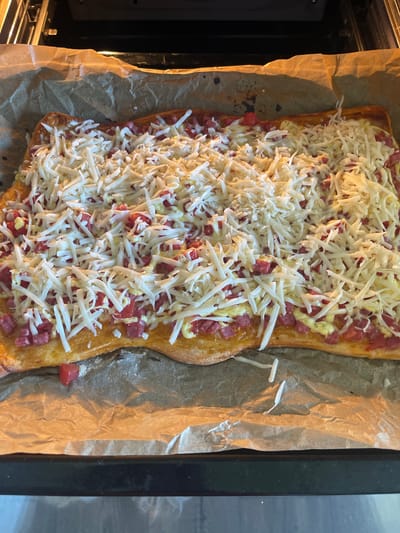 The image depicts a rectangular pizza on parchment paper within an oven. The pizza is topped with tomato sauce, diced pepperoni, and shredded mozzarella cheese. It is located in Verkh-Katunskoye, Russia.