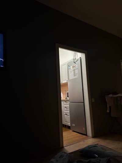 The image depicts an interior scene, likely a residential property in Рублевское Предместье Кп, Russia. The composition is dominated by a doorway leading into a kitchen area, which is partially visible. Inside the kitchen, a silver refrigerator stands against a wall, adorned with various notes and what appear to be magnets. White kitchen cabinets and drawers are also visible, along with a wooden countertop and a faucet. The floor in the kitchen is made of wooden planks.

In the foreground, outside the kitchen, the scene is darker, suggesting a living area. A portion of a television screen is visible on the left edge of the image. On the floor in the foreground, a blue fabric object resembling a blanket or cushion is present, along with some clothing items. A clothes drying rack with laundry is visible in the background, to the right of the doorway. The lighting in the image is dim, indicating either nighttime or a low-light environment. There are no people directly visible in the frame.