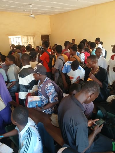 The image displays a densely populated indoor scene, presumably in Ugi Majowel, Nigeria, within a room characterized by light yellow walls and a white ceiling featuring a white ceiling fan. A barred window and a wooden doorframe are visible on the left wall. Approximately 30 to 40 individuals, predominantly young males of African descent, are present, forming a crowded assembly. Most are standing, some with backpacks, and are dressed in various casual clothing including t-shirts and polo shirts. In the immediate foreground, a male in a black shirt is seated on a wooden bench, operating a smartphone. To his left, another male wearing a baseball cap and a patterned shirt holds a blue rectangular object, likely a book or tablet. The overall environment suggests a waiting area, registration point, or a crowded classroom.