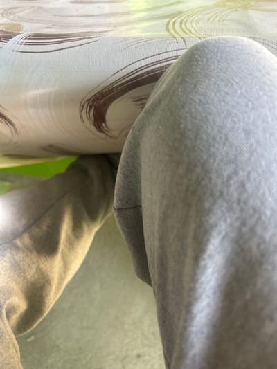 The image depicts a close-up, angled view of a person sitting at a table, likely indoors. The person is wearing light gray sweatpants. The fabric of the pants is visible in detail. A table covered with a patterned, light-colored material is to the left of the pants. The pattern on the table appears to be dark brushstrokes on a lighter background. Behind the pants and table, a section of green and light gray/white surface is visible.
There is insufficient information in the image to determine the location in Kalitinovo, Bulgaria.