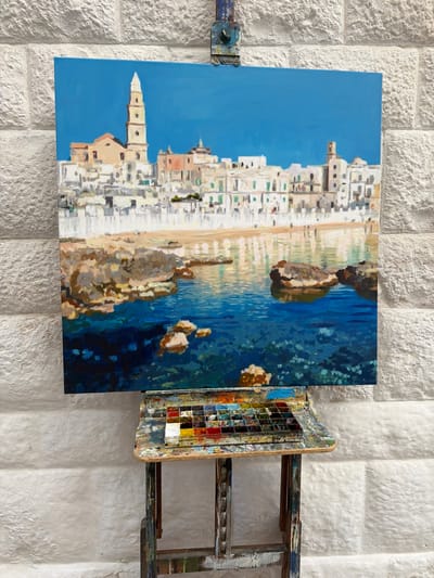 The image shows a painting of Monopoli, Italy, displayed on a wooden easel against a textured white wall. The painting depicts a coastal town with white buildings cascading down to a body of water. The sky is a vibrant blue, suggesting a clear and sunny day. The water is a deep blue, with hints of lighter blues and greens suggesting depth and clarity. Rocks are scattered throughout the water and along the shoreline. The buildings are rendered with a textured brushstroke, giving them a sense of dimension. The painting is framed by the rustic, paint-splattered surface of the easel, which also holds a palette filled with various colors of paint. The overall impression is one of a sunny, Mediterranean scene, likely captured during the daytime. The specific location cues, such as the architectural style of the buildings and the coastal setting, strongly suggest Monopoli, Italy, as indicated by the context. No visible text is present in the image.