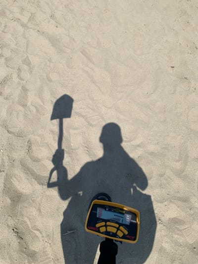 In the city of Indiya, Moldova, a metal detector is being used on a sandy beach. The image captures the shadow of a person holding a shovel, casting a distinct silhouette on the sand. The metal detector, a yellow Garrett Ace 400, is positioned in the foreground, its screen displaying various settings and depth indicators in Russian. Footprints are visible in the sand, suggesting recent activity. The environment is an outdoor, sunlit beach with fine-grained sand. The perspective is from a high angle, looking down at the scene.