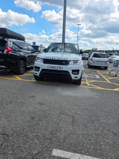 This is a vertical, outdoor, daytime photograph taken in Sholokhovo, Russia. The scene depicts a parking lot with several cars parked in designated spots marked with yellow lines. The foreground is dominated by a white Range Rover, its front end angled towards the viewer. Behind it, a black SUV with a roof box is parked. Further back, other vehicles are visible, including a silver car with a license plate that reads "X186CK 197". The sky is filled with fluffy white clouds against a bright blue backdrop, suggesting fair weather. Tall light poles are scattered throughout the parking lot. The ground is asphalt. Text visible includes the word "ROVER" on the front of the white vehicle, a license plate that reads "O 0999 KE 76 RUS", and a blue parking symbol sign. The overall impression is that of a typical parking scene on a sunny day.
