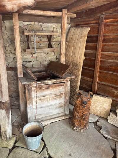 In Zheravna, Bulgaria, an old well is housed within a rustic wooden structure. The well itself is made of weathered wood, with a hinged lid partially open, revealing a dark interior. A metal bucket hangs from a chain attached to the well's structure, positioned on the stone-tiled ground below. To the right of the well stands a carved wooden bear, its features detailed with painted eyes and a black nose. Behind the bear, a carved stone slab with inscriptions leans against the wooden wall of the structure. The walls of the enclosure are constructed from rough stone and rough-hewn timber logs. Various wooden planks and tools are visible within the structure, suggesting it might be a preserved historical exhibit or a functional space from a bygone era. The scene is lit by natural light, casting shadows and highlighting the textures of the aged materials.