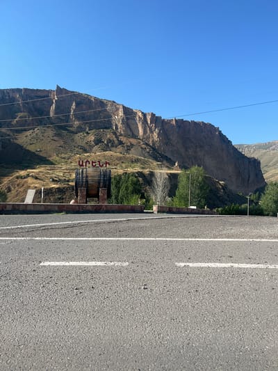 In Zangakatun, Armenia, a large wooden barrel with dark metal bands stands on a stone pedestal in front of a brick wall. Red lettering, in Armenian script, reads "ԱՐԵՆԻ" above the barrel. The barrel and lettering are situated at the base of a sheer, rocky cliff face, with dry, grassy hills rising behind it. A road with white lane markings is in the foreground, and a few trees are visible on the hillside. The sky is a clear, bright blue.