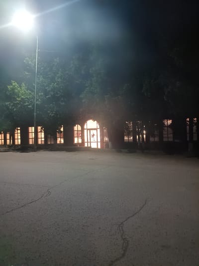 The image depicts a nighttime scene in Ponomarevka, Russia. A bright street lamp illuminates the foreground, casting a hazy glow into the darkness. Behind it, a row of trees with illuminated windows can be seen. The windows are arched and emit a warm, yellow light, suggesting interior illumination. The architecture appears to be a building with multiple arched openings. The ground in front of the building is paved with asphalt, showing visible cracks. The overall atmosphere is dim and somewhat mysterious, characteristic of a quiet night.