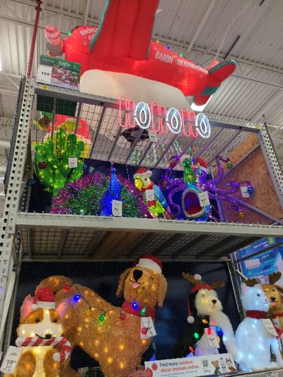 A retail display showcases a variety of Christmas decorations. An inflatable red airplane labeled "Baron the Flying Wor" is next to neon "Ho Ho Ho" lights. A glittery Christmas tree stands near a green decoration, possibly a frog. On the top shelves are lighted Christmas animals, including a reindeer, snowman, and two dogs. The animals are adorned with Santa hats and Christmas lights. Price tags are visible on the decorations. The display structure consists of metal shelving.