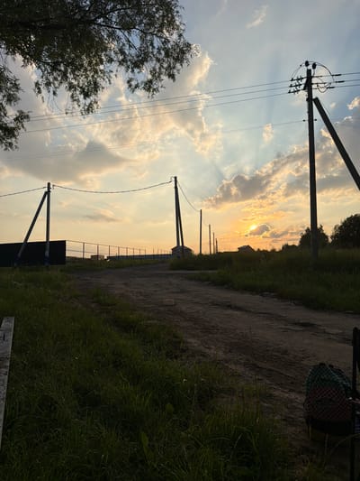 This is a landscape photograph taken in Пропан Снт, Russia, during sunset. The image is oriented vertically, with the horizon running from left to right across the mid-section. A dirt road with grassy verges stretches into the distance, lined with power poles and utility wires. In the background, the sun is setting behind scattered clouds, casting a warm glow across the sky and illuminating the landscape with soft, diffused light. The silhouette of trees and vegetation is visible along the edges of the road. A checkered backpack rests on the ground near the right edge of the frame. The overall mood is peaceful and serene, evoking the end of a day in a rural or semi-rural setting. There is no visible text in the image.
