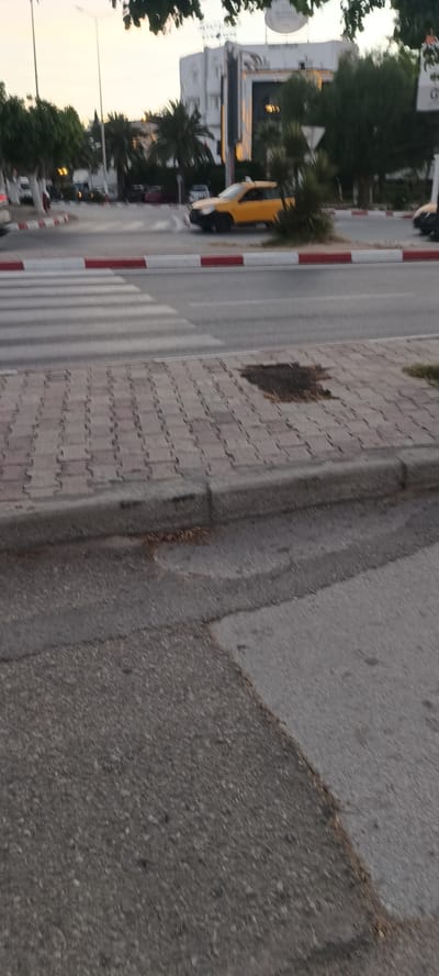 The image captures a street scene in Hammam Sousse, Tunisia, likely during daylight hours given the lighting. The foreground features a paved sidewalk made of interlocking brick pavers, with a curb separating it from the road. A patch of discolored ground, possibly a stain or a missing section of pavers, is visible on the sidewalk. Across the street, a pedestrian crossing with white stripes is laid out on the asphalt road.

In the mid-ground, a yellow taxi is the most prominent vehicle, positioned on the road and facing left. Behind the taxi, trees and palm trees line the street, with a white building visible in the background. The building appears to be a commercial establishment, possibly a hotel or office, with decorative lighting illuminating its facade. The overall atmosphere suggests an urban environment in Hammam Sousse.