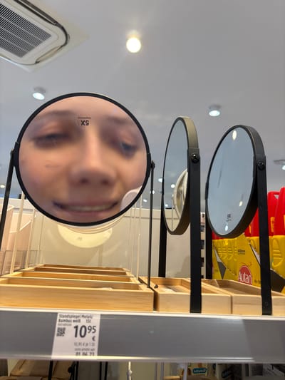 In a retail environment, three round mirrors are displayed on shelves. The leftmost mirror reflects a close-up of a person's face, with a sticker indicating "Vergrößerung 5X" (Magnification 5X). The other two mirrors are less clear in their reflections. Below the mirrors are wooden display trays. A price tag is visible on the shelf, showing "1095" with "Standspiegel Metall/Bambus weiß" (Standing mirror metal/bamboo white) and other details. The overall scene suggests a store, possibly a department store or a specialized retailer. The context of Erlenbach am Main, Germany, is provided, indicating the geographical location of this retail setting.