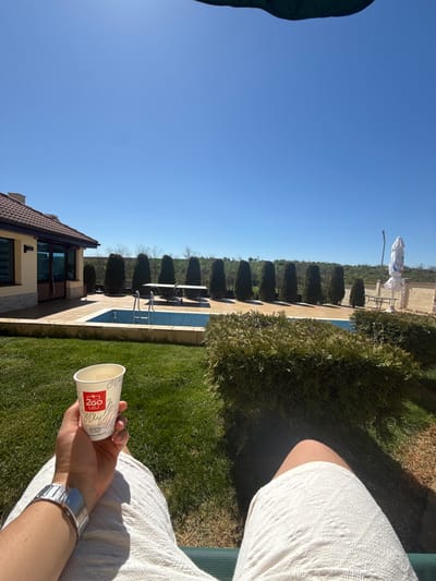 A person sits in an outdoor setting, presumably near the town of Tsar Asen, Bulgaria, based on the "2Go" branding on the paper cup they are holding, which is a Bulgarian coffee chain. The person is wearing a white garment, and a metal wristwatch is visible on their wrist. The foreground includes green grass, and a well-manicured row of trees lines a swimming pool. In the background, there is a building with brown tiled roofing. A clear, blue sky is overhead.