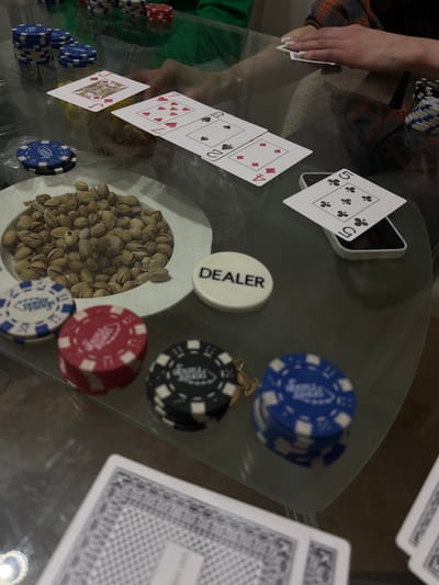 The image captures a casual indoor setting, likely a home, where a card game, possibly poker, is in progress on a glass table. A person's hand is visible on the left, holding a card, wearing a plaid shirt over a green garment.
On the table, several playing cards are laid out: a Five of Clubs standing upright, partially propped against a smartphone; a Four of Diamonds and a Queen of Spades (partially visible) laid flat; a Jack of Diamonds, a Seven of Hearts, and a Ten of Hearts (both partially visible) are also face-up. A stack of face-down cards, likely a deck, is to the right.
Various poker paraphernalia are scattered across the table. Stacks of poker chips in blue, red, and black are visible, some bearing the text "Poker Range." A white, circular "DEALER" button is placed near the center. A white bowl filled with pistachios sits prominently on the table, suggesting a snack during the game. The overall scene conveys a relaxed, social atmosphere. No specific time of day or outdoor weather is discernible, but the lighting suggests an evening or nighttime activity. The location context is Svkh Voronezhskiy, Russia.