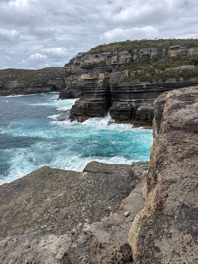 A rugged coastal scene features layered cliffs towering over turbulent ocean waters. The cliffs exhibit a mix of stratified rock formations and green vegetation on top. Waves crash against the cliff bases, creating white foam. A narrow channel of turquoise water is framed between the rock structures, contrasting with the darker, deeper blue of the surrounding sea. The sky is overcast with grey clouds, contributing to the overall dramatic atmosphere.
