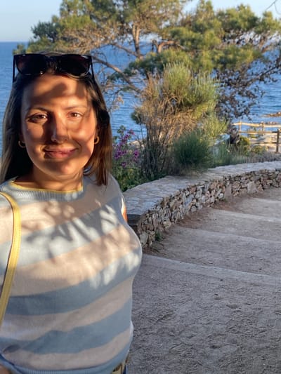 The image depicts a woman in a striped top, wearing sunglasses, standing on stone steps with a stone wall to the right. In the background, there is a view of the sea, trees, and vegetation. The location is Calella De Palafrugell, Spain, given the typical scenery and architectural elements present.
