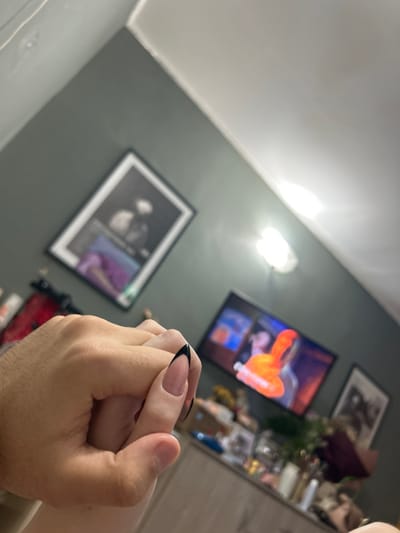An indoor scene is depicted within a residential room in Tsarichino, Bulgaria. The primary focus is on two clasped hands positioned in the lower-middle portion of the frame. The hand on the left is larger, with a light to medium skin tone and short fingernails, appearing to be a male hand. It is positioned over the top of the second hand. The second hand, appearing smaller and with lighter skin, features long, almond-shaped artificial nails with a nude base and black French tips; this is indicative of a female hand. The hands are interlocked or clasped together.
The room features dark grey walls and a white ceiling. On the left wall, a framed black and white picture shows two figures and illegible text below. To the right, a bright, circular ceiling light fixture is visible. A flat-screen television is mounted or placed on furniture in the mid-right, displaying colorful, blurry content, predominantly orange and red. Below the television, a light-colored wooden furniture piece holds various small items, including bottles, containers, and a dark-colored floral arrangement. Another framed picture is visible on the wall to the right of the television.