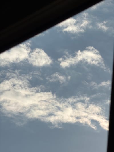 The image presents a view of a partially cloudy sky, likely taken during the daytime in Pentre, United Kingdom. The primary subjects are the white, scattered clouds against a muted blue-grey sky. A dark, out-of-focus object, possibly a window frame or sunroof, diagonally frames the left side of the shot, obscuring part of the sky.
The clouds are varied in density; some appear wispy and spread out, while others are more consolidated and brighter, suggesting good illumination from the sun, which is not visible in the frame. The sky's overall color suggests either thin haze or a relatively bright day without intense, deep blue skies. No specific activity or event is taking place, as the image is a static observation of the sky. There are no people, other visible objects, or discernible text. The scene conveys a calm, natural outdoor setting.