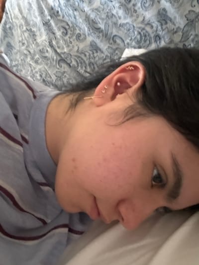 An individual is depicted in a close-up, angled view, lying down on their right side, looking towards the right of the frame. The person has dark hair, visible around the right side of their head. Their right ear is adorned with multiple piercings: a gold-colored hoop in the lobe, two small silver-colored studs above the lobe, a small silver-colored stud in the conch, and a gold-colored, wave-shaped earring in the helix. The visible skin on the face appears fair with scattered minor blemishes. The person is wearing a light blue garment with thin, dark horizontal stripes on the upper torso. The background consists of a light blue and white patterned fabric, likely a blanket or sheet, and a plain white fabric, likely a pillow, supporting the person's head. The scene is set indoors. The specific city of Santiago, Mexico, cannot be determined from the visual information provided in the image.