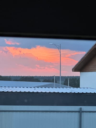 The image captures a view of the sky and rooftops, framed by a dark, rectangular object at the top. The sky is dominated by vibrant pink and orange clouds, suggesting a sunset or sunrise. Two thin, white contrails crisscross the sky, one of which is notably longer than the other. A street lamp stands tall in the middle ground, its pole bisecting the contrails. To the right, the peaked roof of a building with wooden trim is visible, partially obscuring the sky. In the foreground, the corrugated metal roofs of industrial or commercial buildings are prominent. The horizon line features a dense line of dark green trees. The overall scene is an outdoor environment, likely in or near an urban or semi-urban area, given the presence of buildings and a street lamp. The location is specified as Rassokha, Russia.