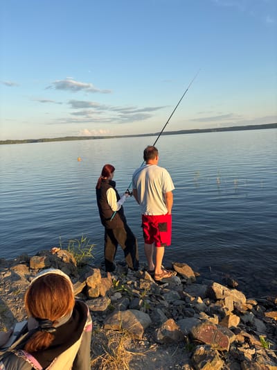 Three people are depicted on the rocky shore of a wide body of water, likely a lake, in Shuiskaya Chupa, Russia. The sky is clear with a few wispy clouds, indicating it is likely late afternoon or early evening. A man in red shorts and a white t-shirt stands facing the water, holding a fishing rod. Beside him, a woman wearing a black vest over a white shirt and black pants also holds a fishing rod, with her hand near the man's. In the foreground, another person, viewed from behind, with reddish-brown hair pulled back in a ponytail, is looking out towards the water. The water is calm and reflects the blue of the sky. A distant, treed shoreline is visible across the water. The foreground is covered in various sized rocks and some sparse vegetation.