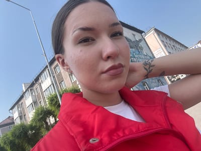 A young woman with dark hair pulled back poses for a selfie outdoors. She is wearing a red vest over a white t-shirt and has a tattoo of a plant branch on her left forearm. Behind her, a multi-story building is visible, with trees and a lamppost in the foreground. The building appears to be in an urban setting, likely in the city of Belovo, Russia, as indicated by the presence of Russian text on a mural on the side of the building. The sky is clear and blue.
