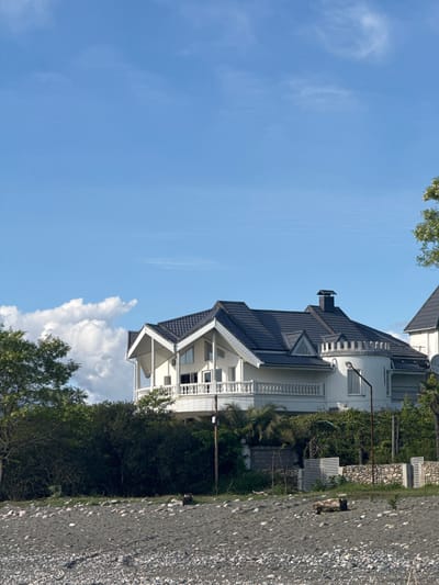 The image depicts a multi-story white house with a dark grey roof located in Gulripshi, Abkhazia, adjacent to a gravel or pebble beach. The house features balconies with white railings, windows, and a chimney. Lush green trees and bushes surround the house. The sky is a bright blue with some scattered white clouds. The foreground shows the beach, which is composed of small rocks. The image seems to be taken during daylight hours.