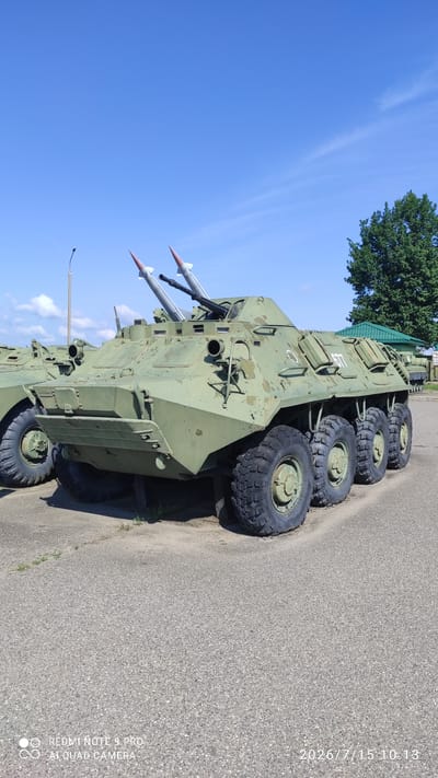 This image shows a green, eight-wheeled military vehicle with missiles mounted on top, parked on an asphalt surface in what appears to be an outdoor display or museum. The vehicle has a weathered paint finish and is seen from a three-quarter front angle. In the background, a tall green tree and a building with a green roof are visible under a clear blue sky with some wispy clouds. Another similar military vehicle is partially visible to the left of the main subject. The overall scene is static and appears to be a sunny day.