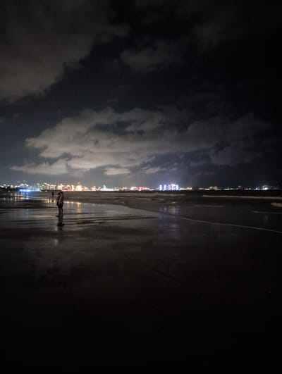 A long shot captures a beach scene at night in Tianya District, China. The sky is dark and cloudy, with faint lights from the distant city visible. The wet sand reflects the lights, creating shimmering patterns. In the foreground, two figures stand close together, silhouetted against the reflective wet sand. The ocean is to the right of the frame, with gentle waves lapping at the shore.