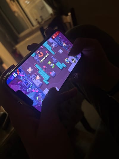 The scene is set indoors in Sofronievo, Bulgaria, under dim lighting conditions. A pair of human hands holds a smartphone horizontally, actively engaged in playing the mobile video game "Brawl Stars." The smartphone screen prominently displays the game's top-down perspective interface, featuring a map with purple ground, blue water-like features, gray structural elements, and various in-game icons. The message "Round lost!" is displayed in white text with a red border near the center of the screen. The individual's thumbs and fingers are positioned over the on-screen controls, with the left hand's thumb near the virtual joystick and the right hand's thumb and index finger near action buttons. A dark bracelet or band is visible on the right wrist. The background is out of focus, showing blurred indoor elements and distant light sources, possibly from a window or other fixtures.