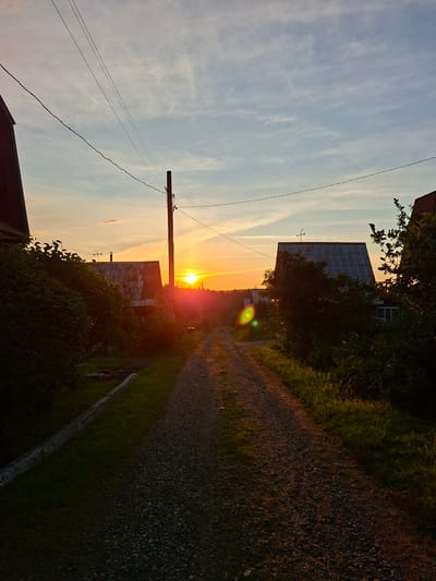 The image depicts a rural road at sunset, likely in a village or dacha settlement near Pervouralsk, Russia. The sun is low on the horizon, casting a warm, golden light across the scene. Power lines stretch across the sky, supported by a wooden pole. On either side of the road, there are houses with metal roofs, partially obscured by trees and foliage. The road itself is unpaved, covered in gravel and dirt. The overall atmosphere is serene and peaceful, with the natural beauty of the sunset dominating the composition.